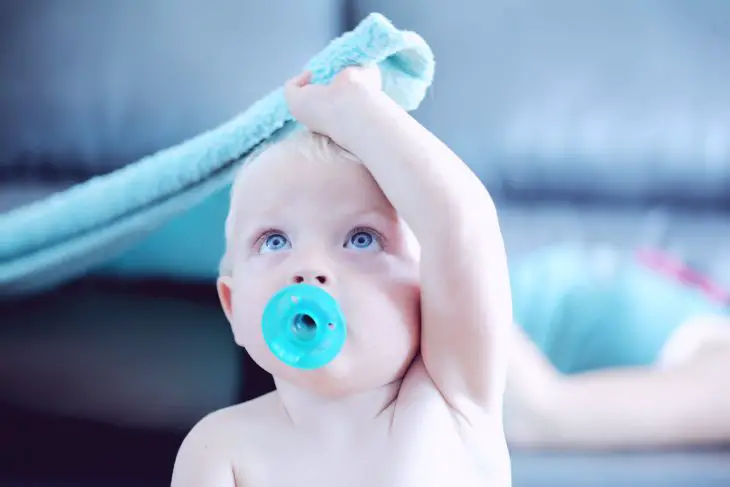 A toddler holding a blue blanket above their head and sucking a pacifier. Ready for nap time!